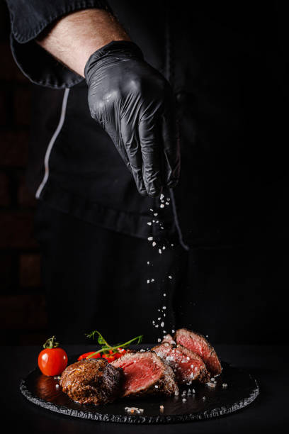 The concept of cooking meat. The chef cook salt on the cooked steak on a black background, a place under the logo for the restaurant menu. food background image, copy space text stock photo