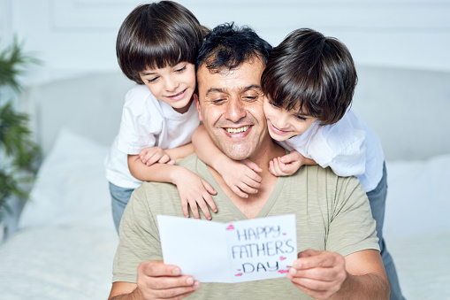 Portrait of latin father looking happy while his two little boys embracing their dad, giving him handmade postcard, greeting with Father's day, spending time together at home. Fatherhood, children