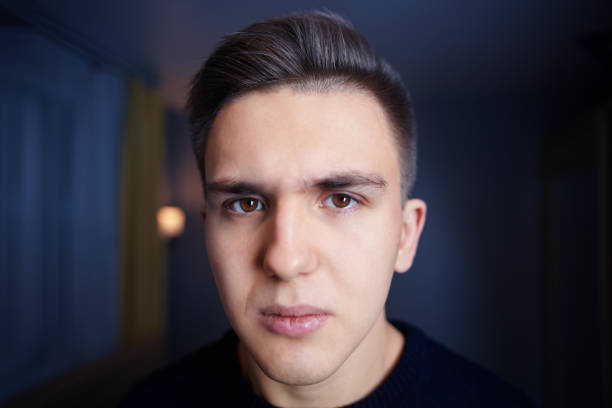 The face of a sullen caucasian guy is 20 years old Close-up the face of a young white man with brown eyes, short hair, and a sullen look, against the background of a dark blue room. Sad Caucasian male 20 years old, in a state of stress, or depression. brown eyes stock pictures, royalty-free photos & images