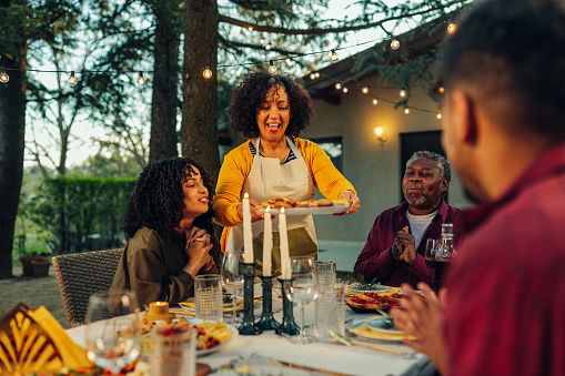 African-American family enjoying dinner outdoors and smiling while sitting at table together, woman brings the food
