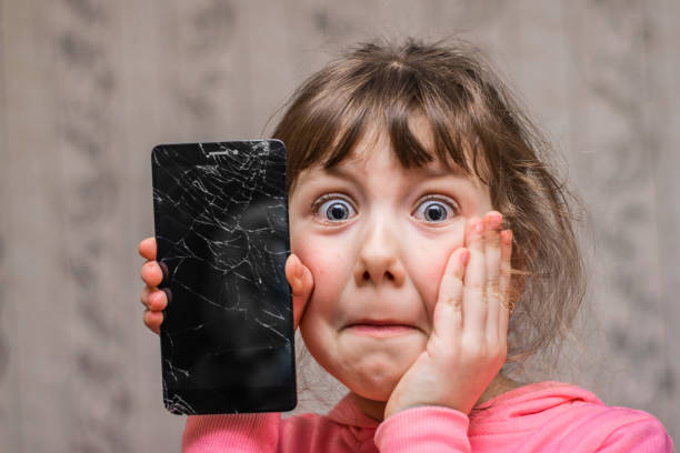 The frightened child accidentally ruined the smartphone. Portrait of a scared little girl with a broken mobile phone. A sad child broke the screen of a mobile phone. Cracked display in hand kids. Portrait of a scared little girl with a broken mobile phone. A sad child broke the screen of a mobile phone. Cracked display in hand kids. broken stock pictures, royalty-free photos & images