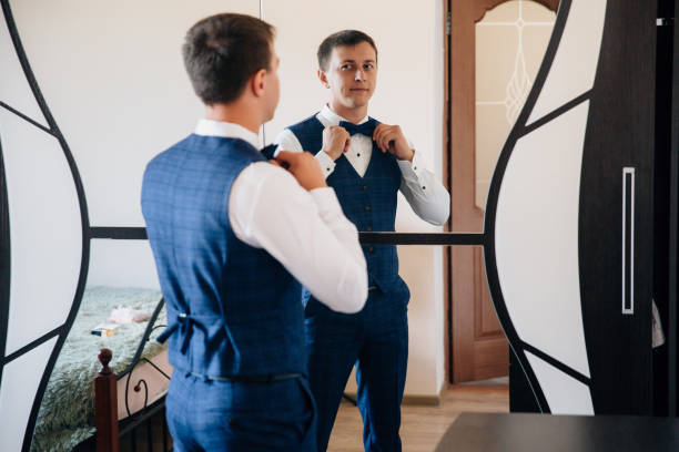 The guy dressed his business suit and tied a stylish bow-tie. The man gathers in front of the mirror and smiles at his reflection stock photo