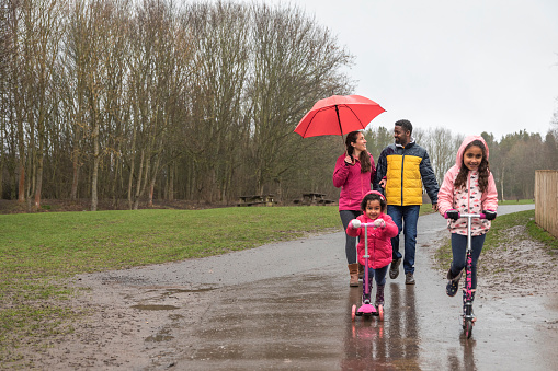 Two young girls riding their push scooters on a wet footpath through Plessey woods, Northumberland. Their parents are walking behind them, the mother is holding an umbrella above her head to shield her from the rain while talking to her husband. They are all enjoying a day out together.