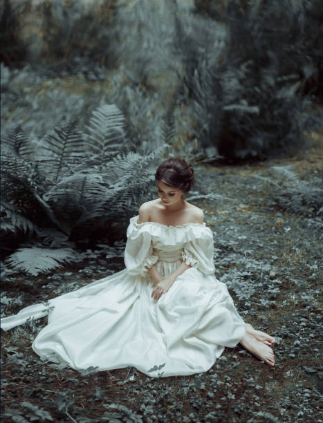 The princess sits on the ground in the forest, among the fern and moss. An unusual face. On the lady is a white vintage dress. Artistic photography. Emotions of melancholy and depression. Cold toning The princess sits on the ground in the forest, among the fern and moss. An unusual face. On the lady is a white vintage dress. Artistic photography. Emotions of melancholy and depression. Cold toning. victorian gown stock pictures, royalty-free photos & images