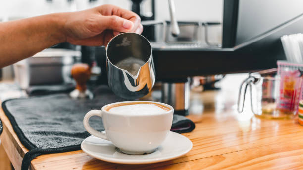 The professional barista pours the milk in the stainless steel mug into the hot coffee cup to make a cappuccino or latte in the coffee shop. how to make cappuccino coffee "n stock photo