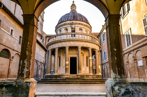 A suggestive perspective of the front side of the Tempietto in the courtyard of the church of San Pietro in Montorio, located on the Janiculum hill in the historic district of Trastevere, in the heart of Rome. Built in 1510 by the architect Donato Bramante, the Tempietto (Small Temple) is considered a masterpiece of Renaissance architecture, with the search for a perfect balance of shapes and volumes. The circular plan and the relationship between the dome and the colonnade recalls the classical values of Roman architecture, such as the famous Temple of Vesta located in the Roman Forum. According to tradition, the Tempietto was built to celebrate the martyrdom of St. Peter, which supposedly took place on the Janiculum. Trastevere is an iconic district of the Eternal City, due to the presence of countless artistic and historical treasures, monuments and ancient Romanesque and Baroque churches, but also for its squares and alleys to be explored freely, where it is easy to find typical restaurants, pubs, small shops of artisans and scenes of daily life with the original Roman soul. In 1980 the historic center of Rome was declared a World Heritage Site by Unesco. Super wide angle and high definition image.