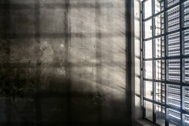 The very sober interior of a prison cell: barred windows with little light coming in and bare concrete walls stock photo