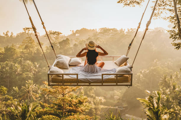 The view is best from above Photo of a young woman sitting on the swing, holding her hat.  Jungle Bed hanging over the tropical forest with Caucasian female resting while looking at the view, Bali, Indonesia. bali stock pictures, royalty-free photos & images