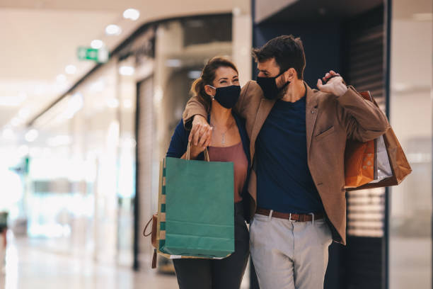 la joven pareja lleva bolsas de compras y camina por el centro comercial, usando máscaras protectoras, la vida en un momento de pandemia - ir de compras fotografías e imágenes de stock