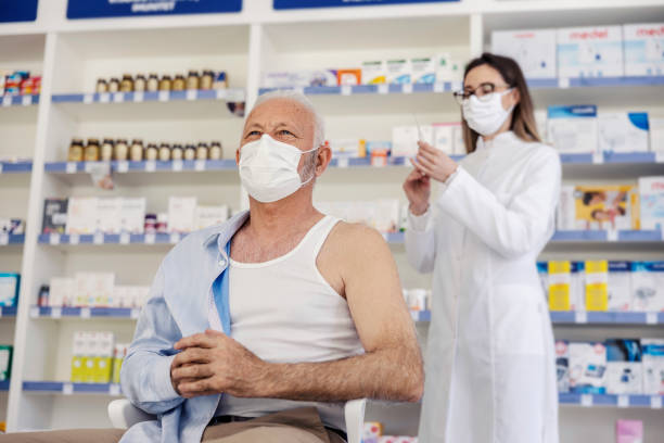 Therapy in a nursing home pharmacy. Female pharmacist gives therapy to a senior man who is sitting on a chair and has taken off his shirt. Vaccination, corona virus breaking news stock photo