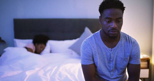 Things don't feel the same anymore Shot of a concerned looking young man sitting on a bed while his wife sleeps in the background insomnia stock pictures, royalty-free photos & images