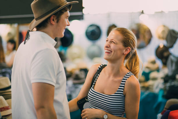 I Think This One Suits You Best! Young couple are exploring Queen Victoria Market in Australia. The woman has put a hat on her boyfriend in a market stall. queen victoria market stock pictures, royalty-free photos & images