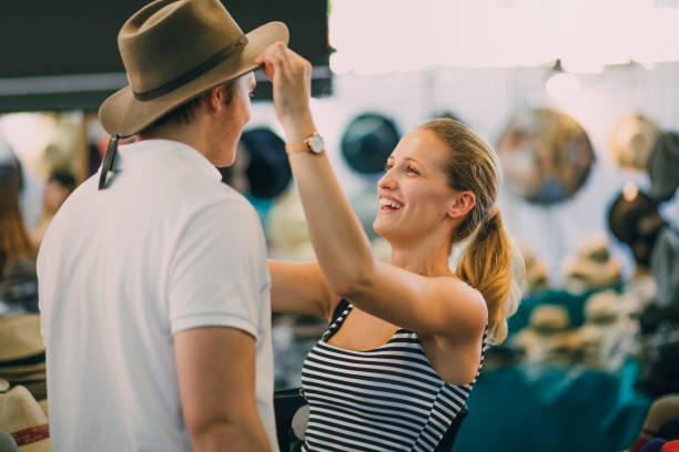 I Think This One Suits You Best! Young couple are exploring Queen Victoria Market in Australia. The woman is putting a hat on her boyfriend in a market stall. queen victoria market stock pictures, royalty-free photos & images