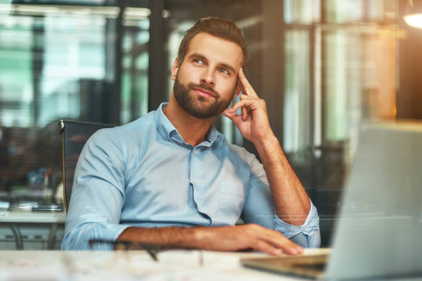 Thinking about business. Young bearded man in formal wear touching his head, smiling and looking away while working in modern office stock photo