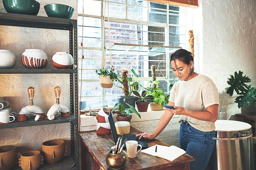 Cropped shot of an attractive young business owner standing and using her laptop and cellphone in her pottery studio