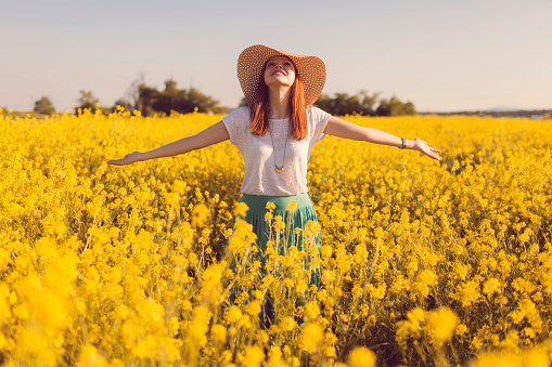 Beautiful, russet young woman with a hat standing in the middle of the flower meadow, surrounded by yellow flowers, with her arms outstretched