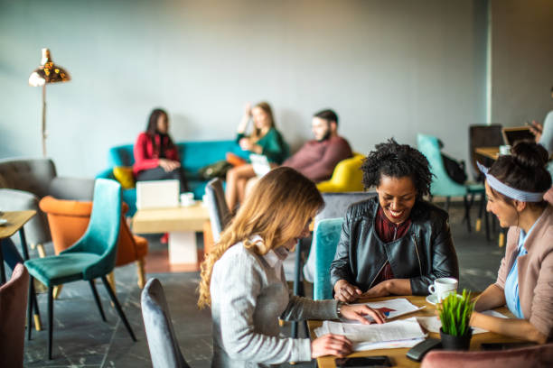 This is the perfect spot for coffee and handle business Multi-ethnic group of female colleagues sitting and looking at paper document in cafe on coffee break coworking stock pictures, royalty-free photos & images
