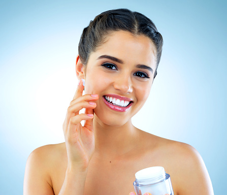 Studio shot of a beautiful young woman applying moisturizer to her face against a blue background