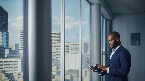 Thoughtful Black Businessman in a Tailored Suit Using Laptop while Standing in Office Near Window on Big City. Successful Corporate Top Manager Doing Data Analysis for e-Commerce Startup
