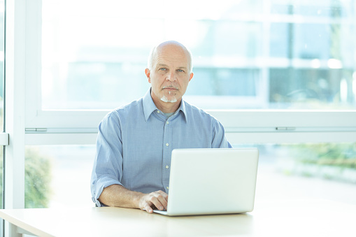 Thoughtful businessman sitting working at his laptop computer in a bright high key office against a window backdrop staring intently at the camera with deadpan expression