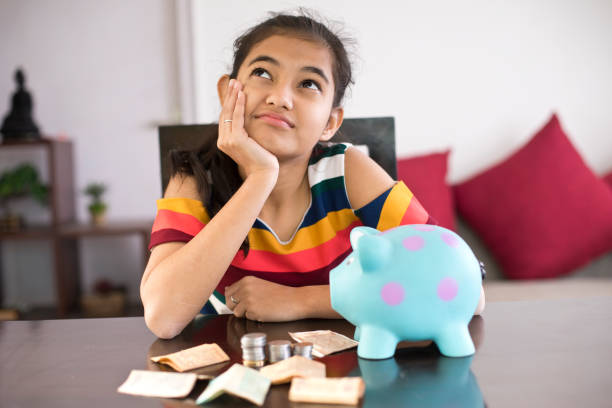 Thoughtful girl with her savings Girl day dreaming with piggy bank and money on table money stock pictures, royalty-free photos & images