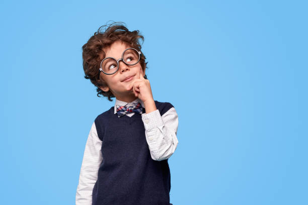 Thoughtful little genius looking up Smart wunderkind in nerdy glasses and school uniform touching cheek and looking up while thinking against blue background boys glasses stock pictures, royalty-free photos & images