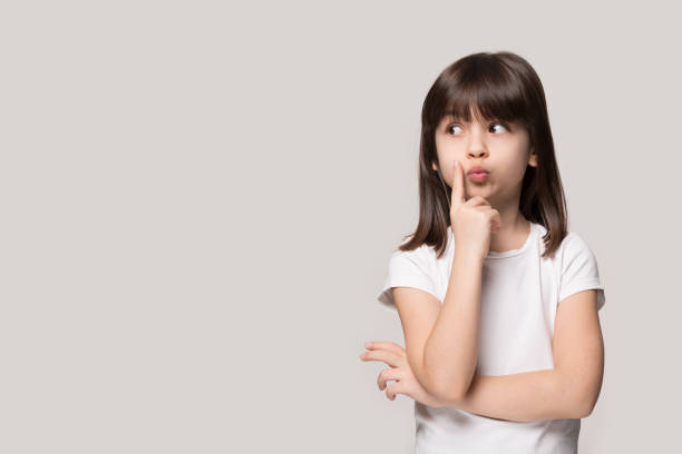 Thoughtful little girl looking away thinking isolated on grey background Thoughtful little girl six years old daughter look away on copy space thinking isolated on grey beige background, funny kid pout lips hold finger near mouth conceived some kind of prank, concept image child behaving badly stock pictures, royalty-free photos & images