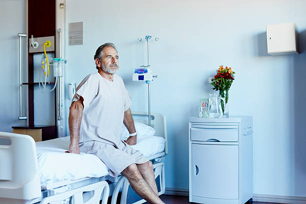 Thoughtful mature man in hospital ward Thoughtful mature man looking away while sitting on bed in hospital ward patient in hospital bed stock pictures, royalty-free photos & images