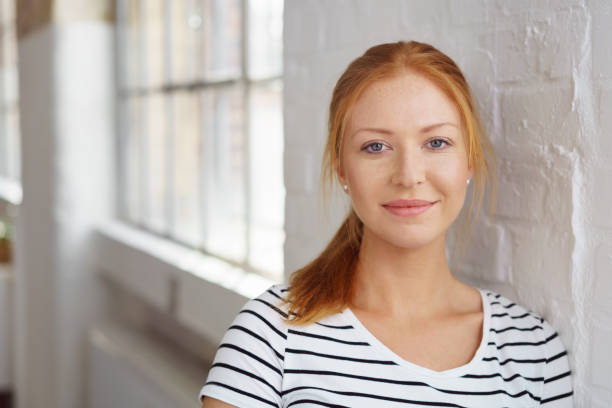 Thoughtful pretty woman leaning on a wall Thoughtful pretty young redhead woman in a striped t-shirt leaning on a white painted brick interior wall looking at the camera with a quiet smile young women stock pictures, royalty-free photos & images