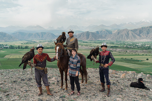 Three eagle hunters, one of them on horse,  and female traveler standing in steppe in Kyrgyzstan