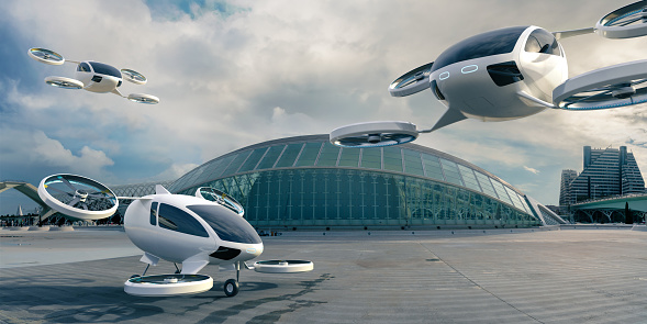 A fleet of generic white electric powered Vertical Take Off and Landing eVTOL aircraft with four rotors. One aircraft is parked with landing gear on empty concrete surface in front of glass terminal building on an overcast day. Two other eVTOL's are in mid flight in the background and foreground.