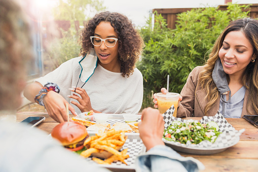 Three female friends enjoy lunch together at an outside table during the coronavirus outbreak.  They partially remove their masks in order to eat.