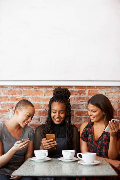 Three Female Friends Meeting For Coffee Sitting At Table Looking At Mobile Phones Three Female Friends Meeting For Coffee Sitting At Table Looking At Mobile Phones fomo photos stock pictures, royalty-free photos & images