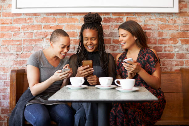 Three Female Friends Meeting For Coffee Sitting At Table Looking At Mobile Phones Three Female Friends Meeting For Coffee Sitting At Table Looking At Mobile Phones fomo photos stock pictures, royalty-free photos & images