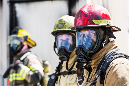 A group of three multiracial firefighters wearing protective gear, including an oxygen mask, helmets, and tan protective suit with yellow reflective stripes.  The focus is on the fireman on the right, a mixed race African American man wearing a red helmet.  The one in the background is a woman.