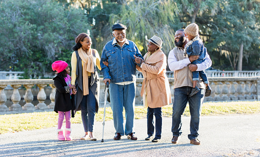 A multi-generation African-American family taking a walk together on a sunny autumn day at the park. The grandfather is in his 70s walking with a cane and being supported by grandma on one arm and his daughter on the other. The granddaughter is 6 years old, and the grandson, in dad's arms, is 3.