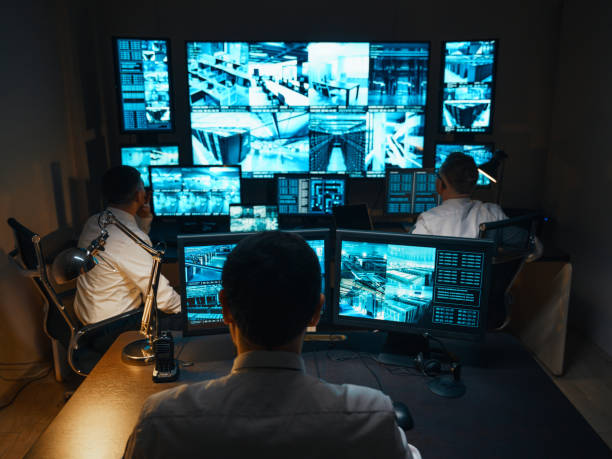 Three guards are in the security room. They sit at work stations with large monitors that display real-time video images from security cameras. The guards are ready for any eventuality. stock photo