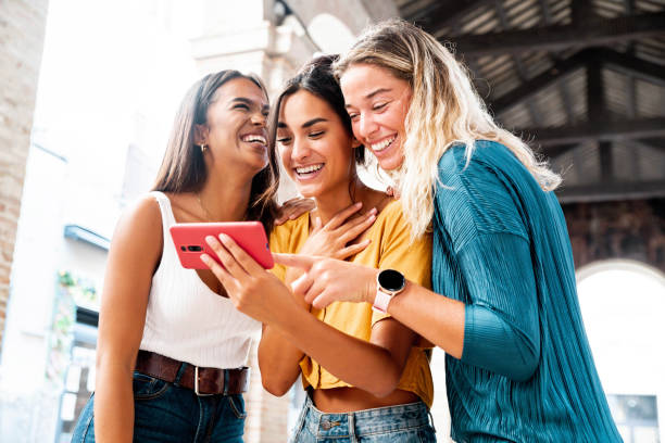 Three happy friends watching a smart phone mobile outdoors - Millennials women using cellphone on city street - Technology, social, friendship and youth concept stock photo