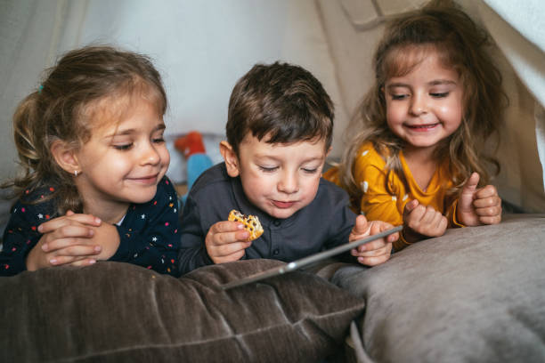 Three kids and digital tablet Portrait of three little kids at home, lying in tent, eating cookies and watching digital tablet fort stock pictures, royalty-free photos & images