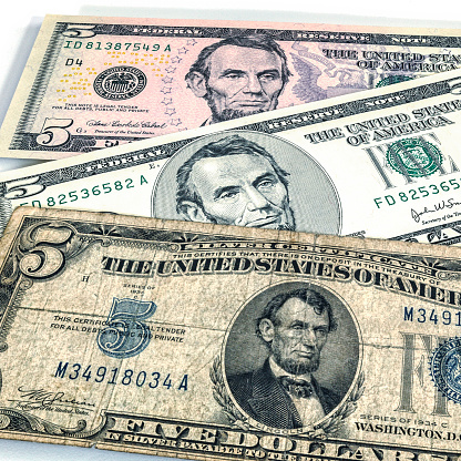 Group of three United States five dollar paper currency bills of significantly different eras, designs and series. In the foreground, at the bottom, is a well-worn, wrinkled, distressed, torn and damaged vintage 1930's era Silver Certificate. In the middle is the surprisingly bland - by comparison to the others - late 20th-early 21st century five dollar Federal Reserve Note. In the background at the top is an example of the most current (as of 2018) colorful, complicated, security-enhanced Federal Reserve Note five dollar bill.