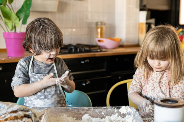 Three year old boy looking with surprised expression at his hands covered with flour, while his little girl concentrates on making the dough stock photo