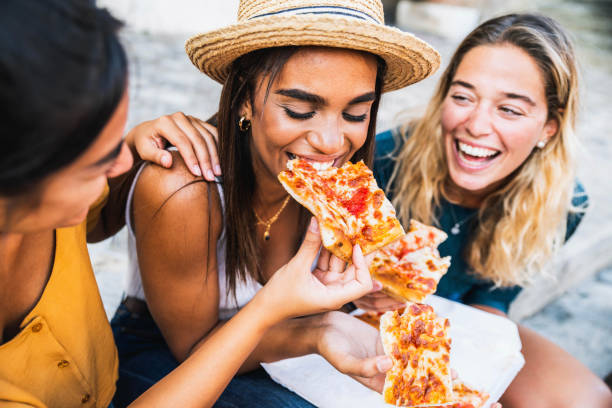 Three young female friends sitting outdoor and eating pizza - Happy women having fun enjoying a day out on city street - Happy lifestyle concept stock photo