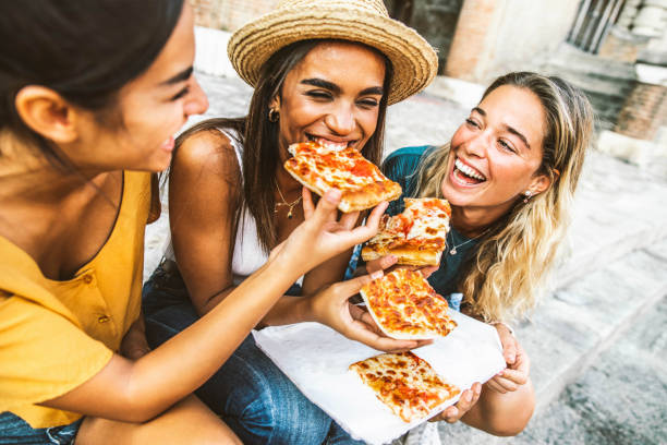 Three young female friends sitting outdoor and eating pizza - Happy women having fun enjoying a day out on city street - Happy lifestyle and tourism concept stock photo