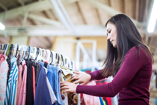 Thrift Store Shopping Young Woman A beautiful young adult woman in her early 20's shops at a second hand thrift shop, searching through racks of clothes for cheap finds and treasures.  She looks through a rack of clothes for something that will fit.  Horizontal with copy space. thrift store photos stock pictures, royalty-free photos & images