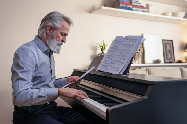 Tidy gray-haired, bearded, charming composer, pianist, middle-aged artist in a blue shirt at home in a brightly decorated room, concentrated stacks and plays the electric piano and stacks tunes with the help of wireless headphones. stock photo
