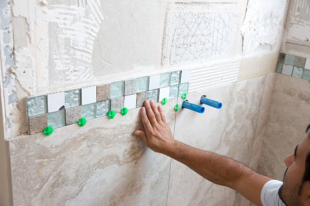 Tile series:Tile border being installed on shower wall in home Tile border made up of small square tiles in glass, metal and stone are being adhered to the wall of a shower. Larger tiles are in place underneath it. The bathroom in a home The arm and head of tile worker are visible.  Taken with a Canon 5D Mark 3.  rm bathroom walls stock pictures, royalty-free photos & images