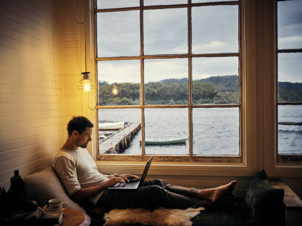 Time away from the world...or not quite Shot of a young man using a laptop while relaxing in his holiday home airbnb stock pictures, royalty-free photos & images