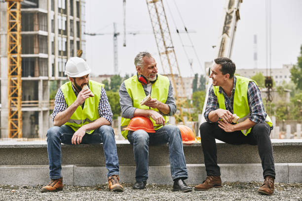 休息的時間到了。一群穿著工作服的建築工人在坐在石頭表面對建築工地的時候吃三明治和說話。建築理念。午餐概念 - construction worker 個照片及圖片檔