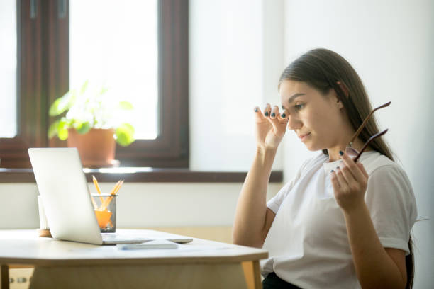 Tired businesswoman holding glasses and rubbing eyes in home office Young female worker with tired eyes holding glasses. Woman feeling discomfort from long wearing eyeglasses behind laptop at workplace. Eyesight strain from computer work concept staring stock pictures, royalty-free photos & images