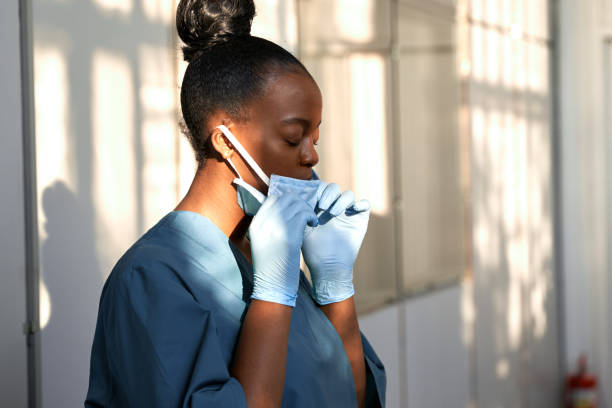 Tired female african scrub nurse wear blue uniform gloves taking off face mask to breathe in hospital. Exhausted black doctor feels stress relief concept, burnout fatigue at difficult work in hallway. Tired female african scrub nurse wear blue uniform gloves taking off face mask to breathe in hospital. Exhausted black doctor feels stress relief concept, burnout fatigue at difficult work in hallway. frontline worker stock pictures, royalty-free photos & images